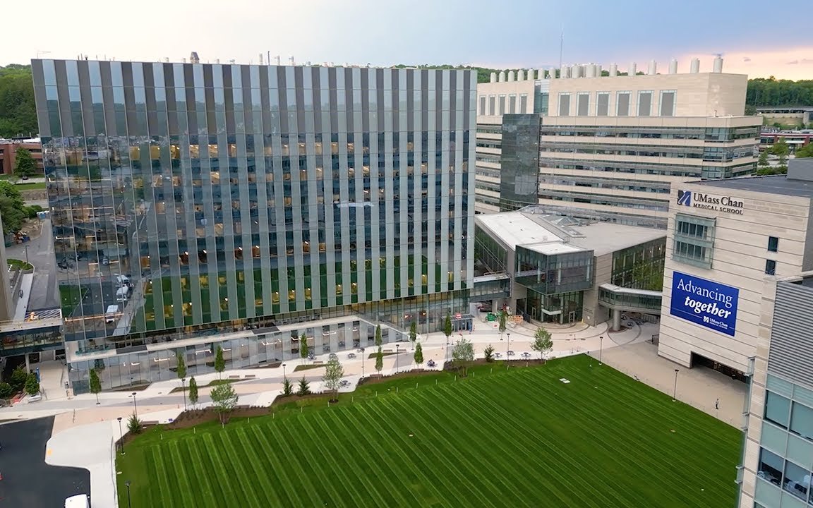 Aerial view of the UMass Chan campus: a tall glass-clad building, the Paul J. DiMare Center, beside stone research buildings and a wide green lawn